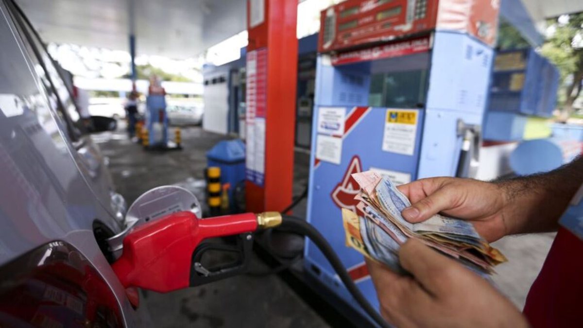 Um homem abastece um veículo enquanto segura notas de dinheiro - Agência Brasil - Preços abuvisos postos de combustíveis