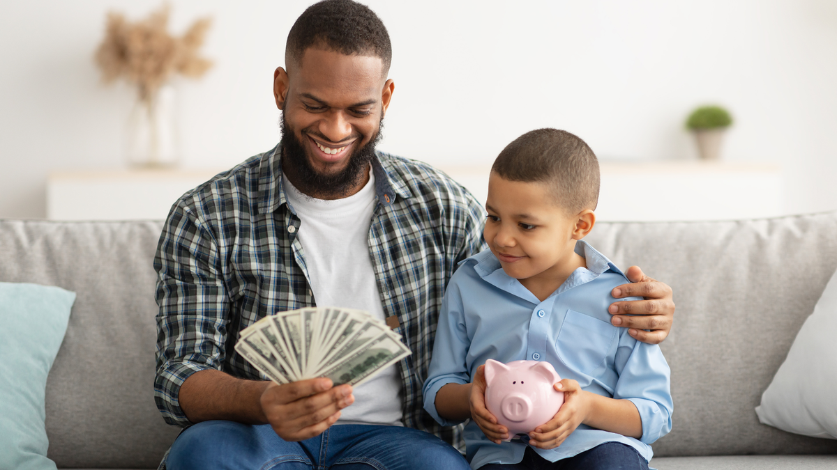 Pai abraça filho e segura notas de dinheiro enquanto criança segura cofrinho em formato de porco - Divulgação
