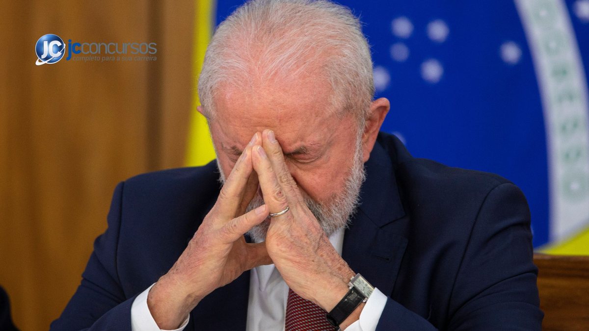 Presidente Lula com cabeça baixa e mãos no rosto e a bandeira do Brasil ao fundo