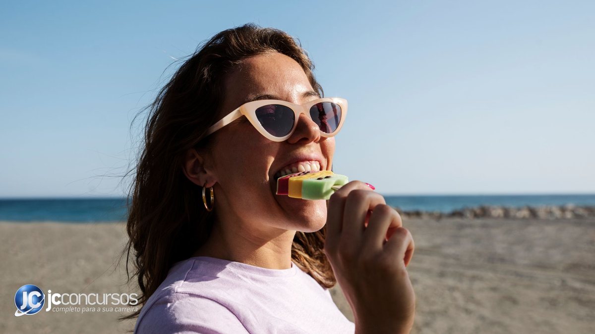 Mulher tomando picolé na praia enquanto usa óculos escuros - Freepik