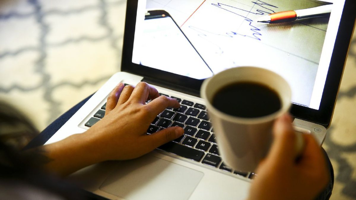Pessoa segura caneca com café enquanto digitam em teclado de notebook